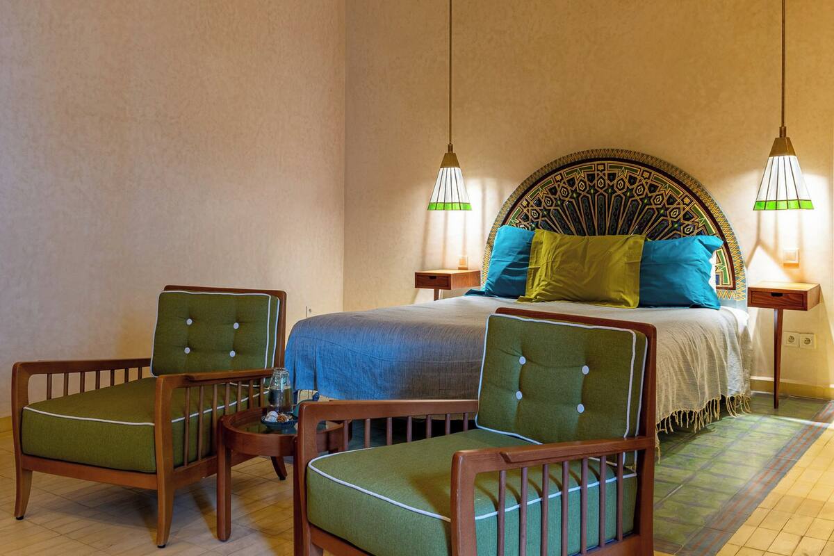 A hotel room with a bed, two chairs, a nightstand, and two wall-mounted lamps.