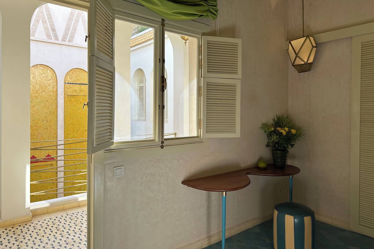 A room with a window, a small table, a potted plant, and a wall-mounted light fixture.