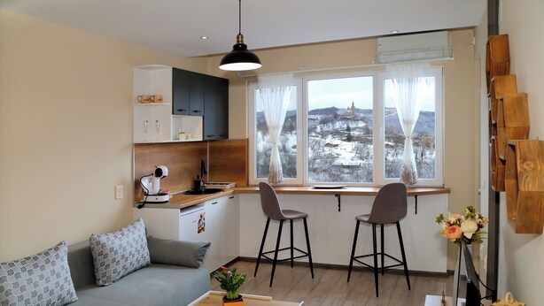 A modern living room with a grey sofa, a wooden coffee table, and a view of a snowy village through large windows. - Colibri Castle View – Romantic Escape (Veliko Tarnovo)