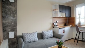 A modern living room with a grey sofa, a wooden coffee table with a plant, and a kitchen area with white cabinets and a grey chair. - Colibri Castle View – Romantic Escape (Veliko Tarnovo)