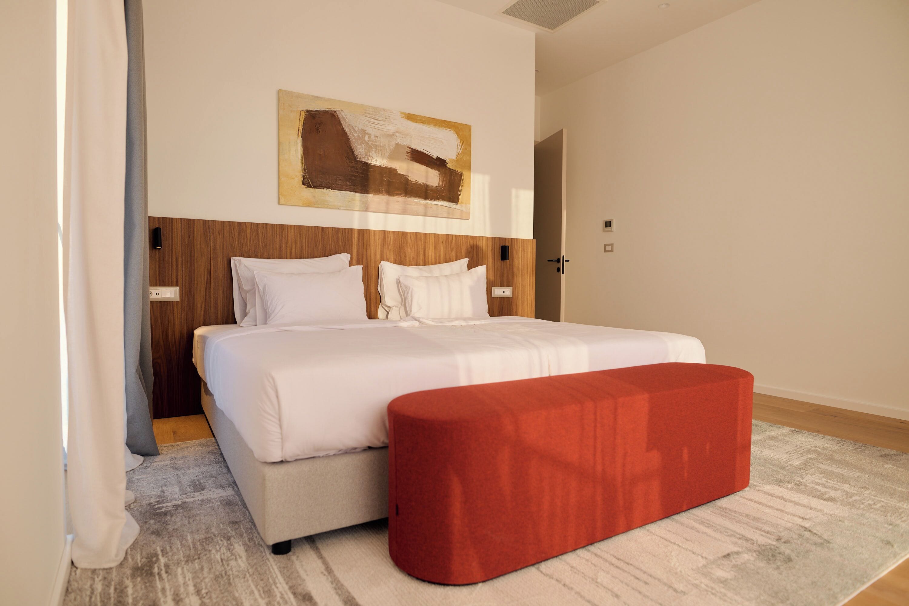 A modern hotel room with a large bed, a red ottoman, and a painting on the wall.