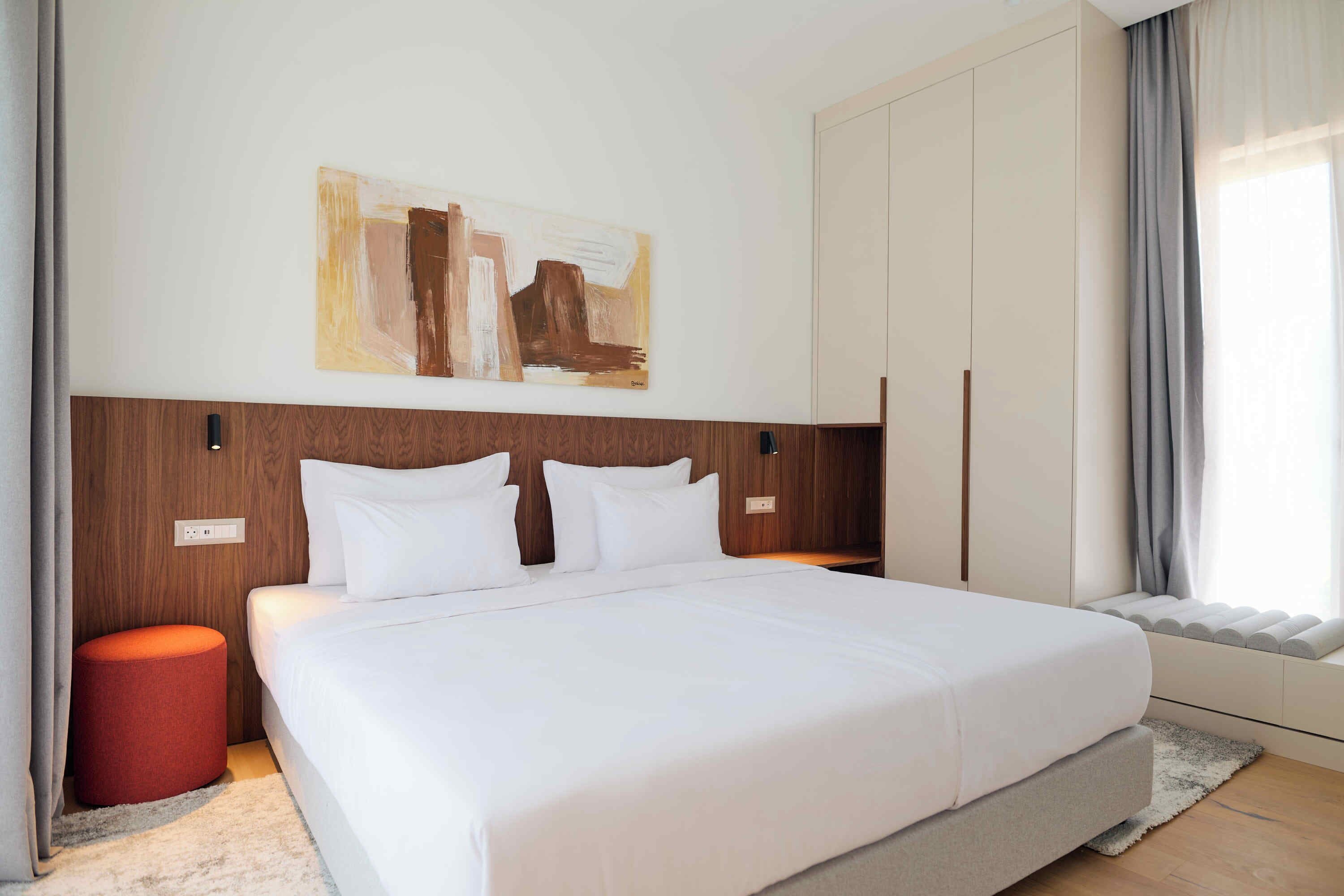 Una camera d'albergo moderna con un letto grande, una testiera in legno, un poggiapiedi rosso, un quadro appeso al muro e una finestra con tende.