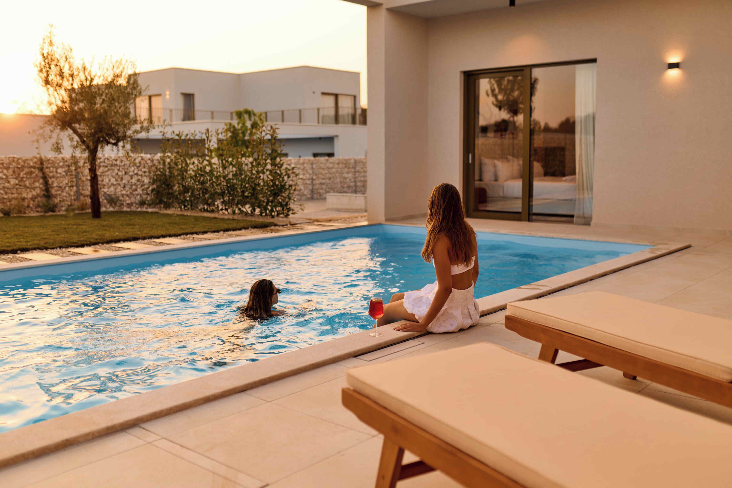 A pool area with a person sitting on a lounge chair, a dog in the water, and a building in the background.