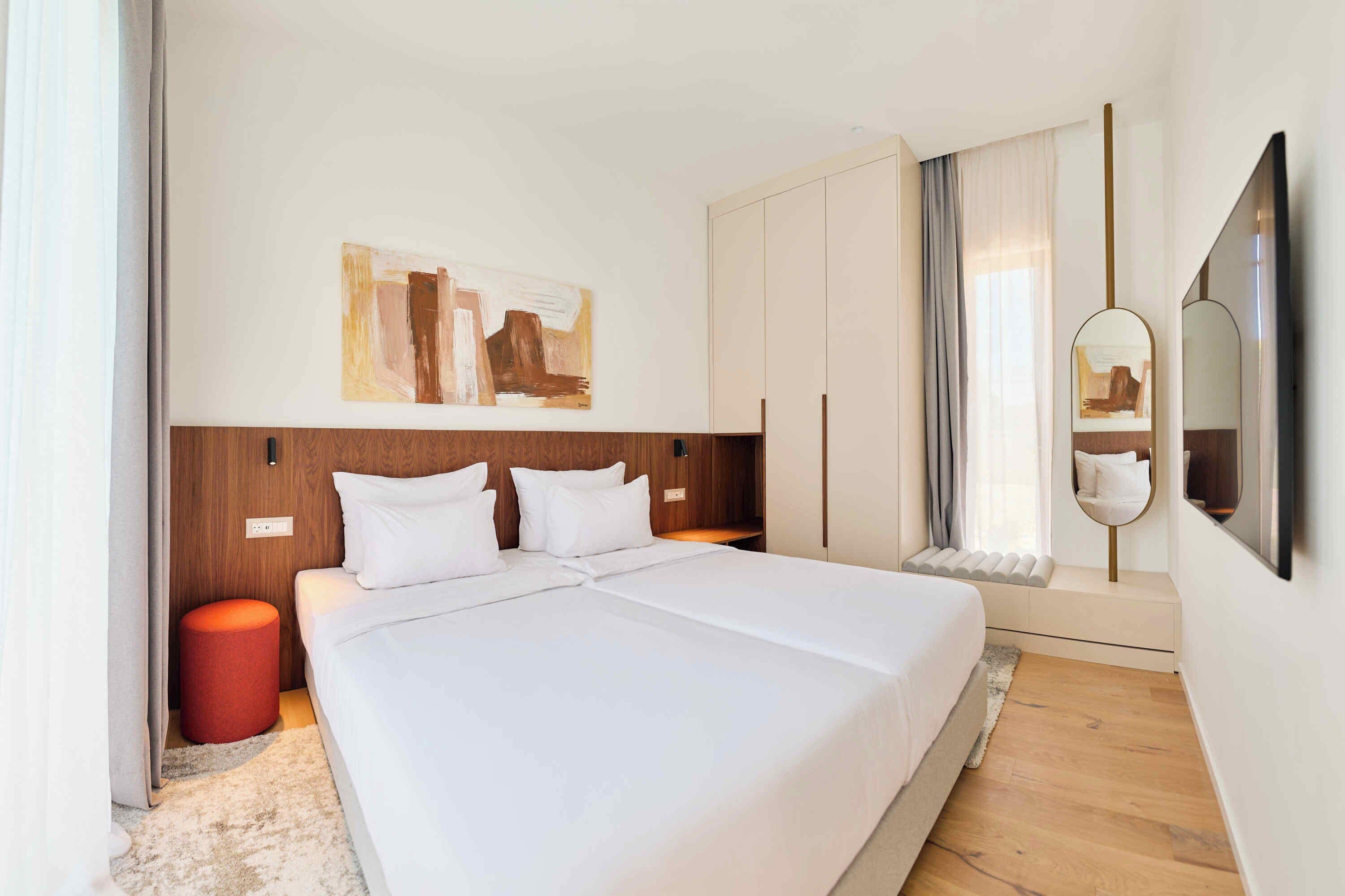 A hotel room with a large bed, a wooden headboard, a painting on the wall, a wardrobe, a mirror, and a window with curtains.