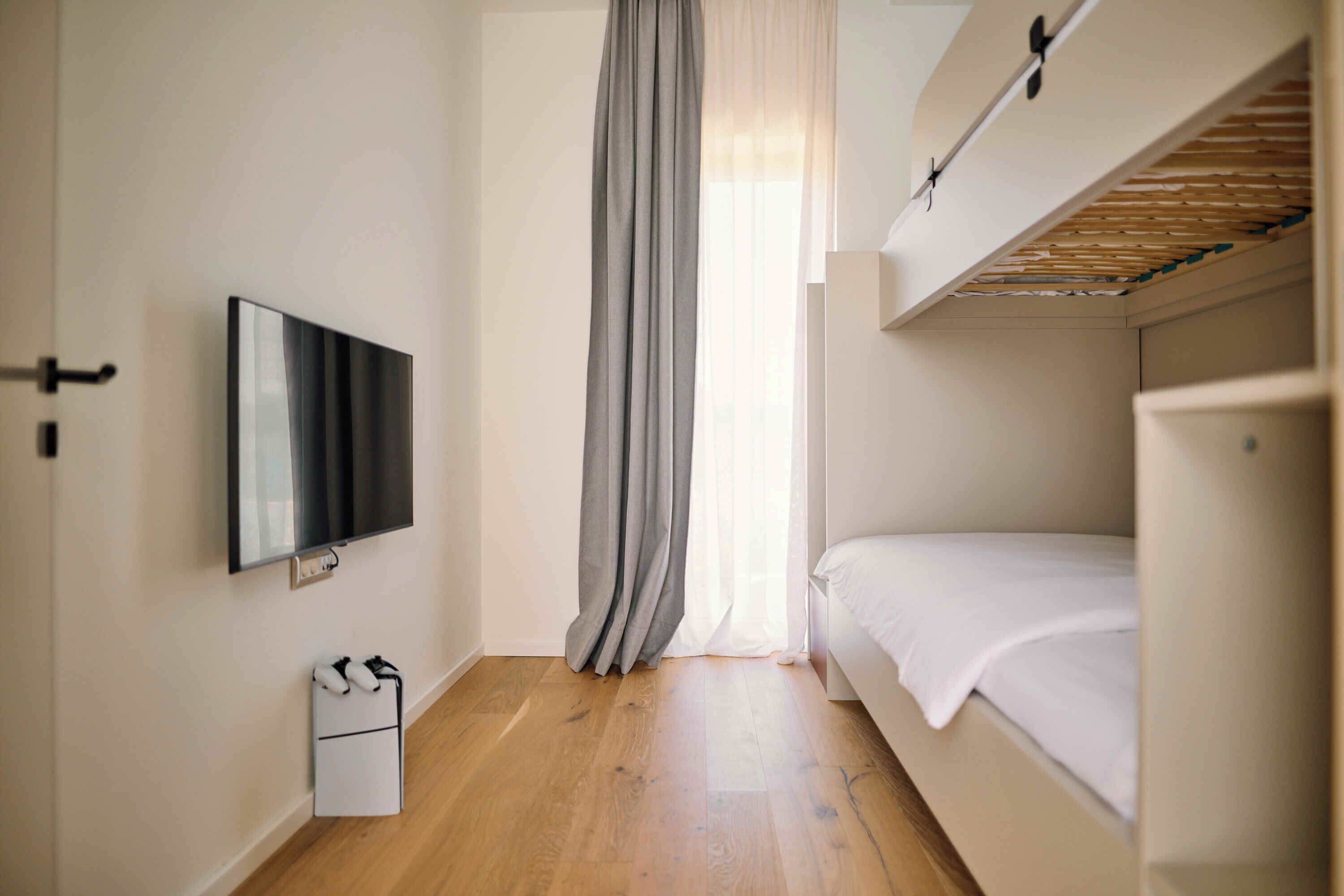 Una habitación con una cama, un televisor montado en la pared, suelo de madera y una ventana con cortinas.