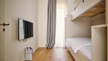 Una habitación con una cama, un televisor montado en la pared, suelo de madera y una ventana con cortinas.