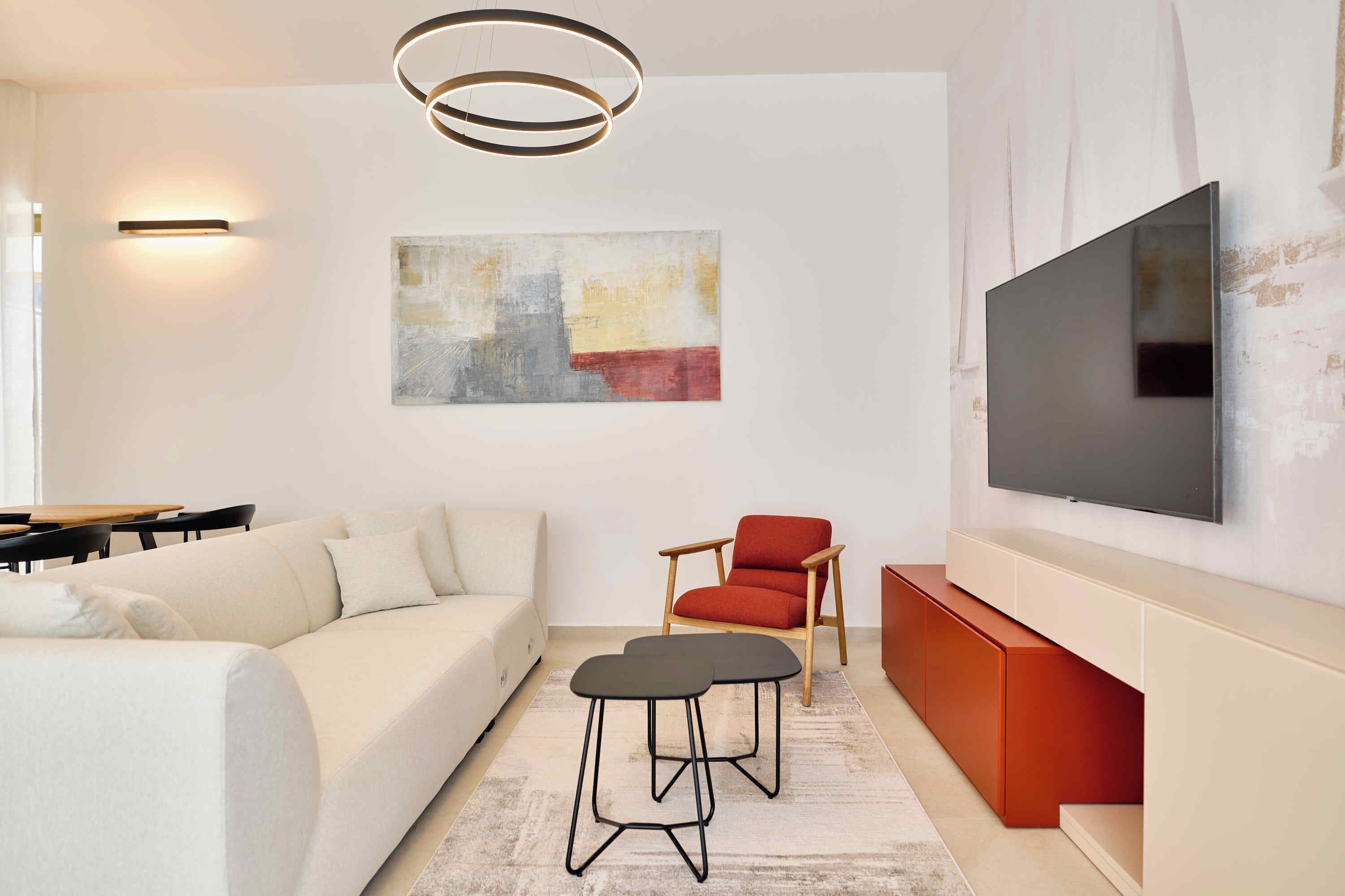 A modern living room with a sofa, two black stools, a red chair, a flat-screen TV, and abstract art on the wall.