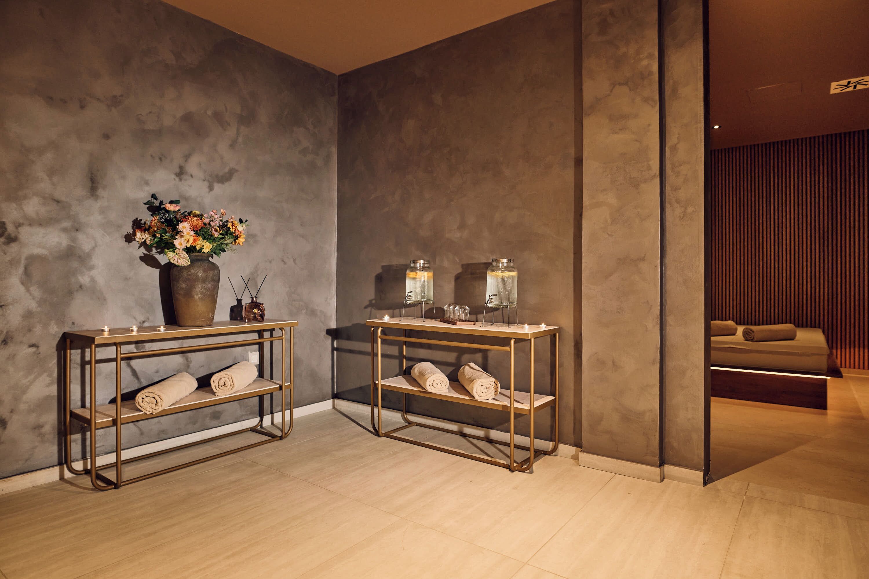Une salle de spa moderne avec deux tables aux cadres dorés, chacune agrémentée d’un vase de fleurs et de bougies, ainsi qu’un banc en bois sur lequel sont posées des serviettes.