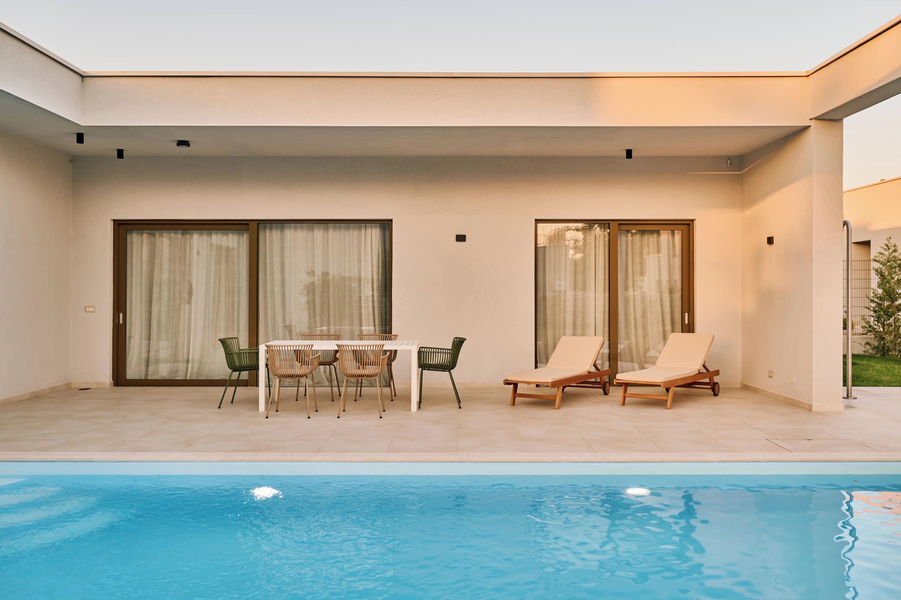 Un área de piscina moderna al aire libre con una mesa y sillas, dos sillones de descanso y un patio cubierto con puertas de vidrio correderas.