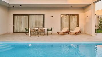 Un área de piscina moderna al aire libre con una mesa y sillas, dos sillones de descanso y un patio cubierto con puertas de vidrio correderas.