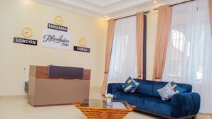 A living room with a blue sofa, a glass coffee table, and a reception desk. There are clocks on the wall indicating different cities. - Plavender inn (Dar es Salaam)