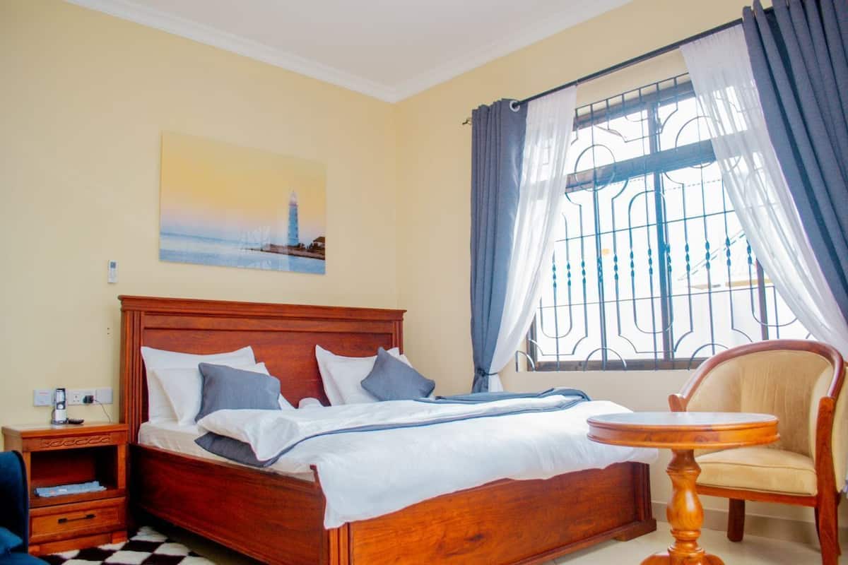Une chambre avec un lit en bois, une table de chevet, une chaise, une fenêtre avec des barreaux en métal et un tableau accroché au mur.