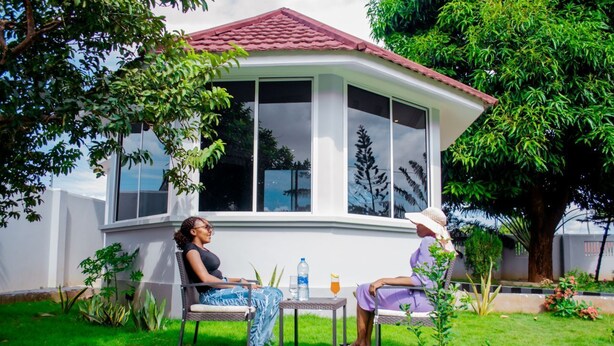 A covered patio with two chairs and a table, surrounded by greenery and a building with large windows. - Plavender inn (Dar es Salaam)