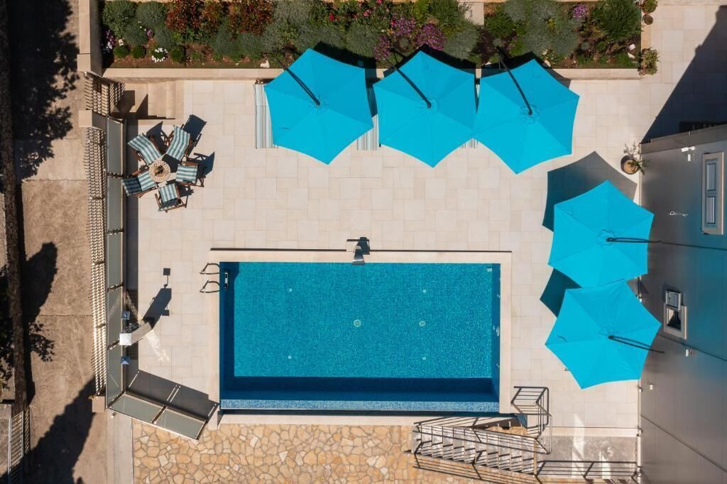 Vista aérea de un área de piscina con varios sombrillas azules, sillas de descanso y un suelo de baldosas de piedra.