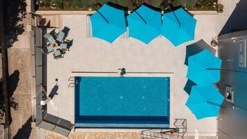 Vista aérea de un área de piscina con varios sombrillas azules, sillas de descanso y un suelo de baldosas de piedra.