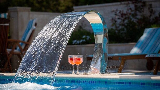 Una piscina con elemento d'acqua, due lettini prendisole e un bicchiere con una bevanda.
