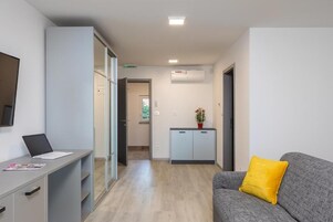 Una habitación de hotel moderna con un escritorio, un sofá gris con un cojín amarillo, un mueble bajo con una planta y una puerta de vidrio corredera que da a otra habitación.
