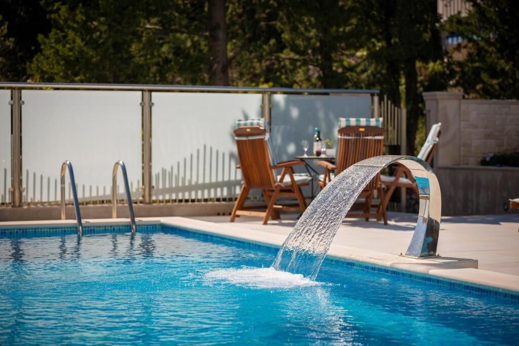 A swimming pool with a water feature, surrounded by wooden chairs and a table, with a fence and trees in the background.