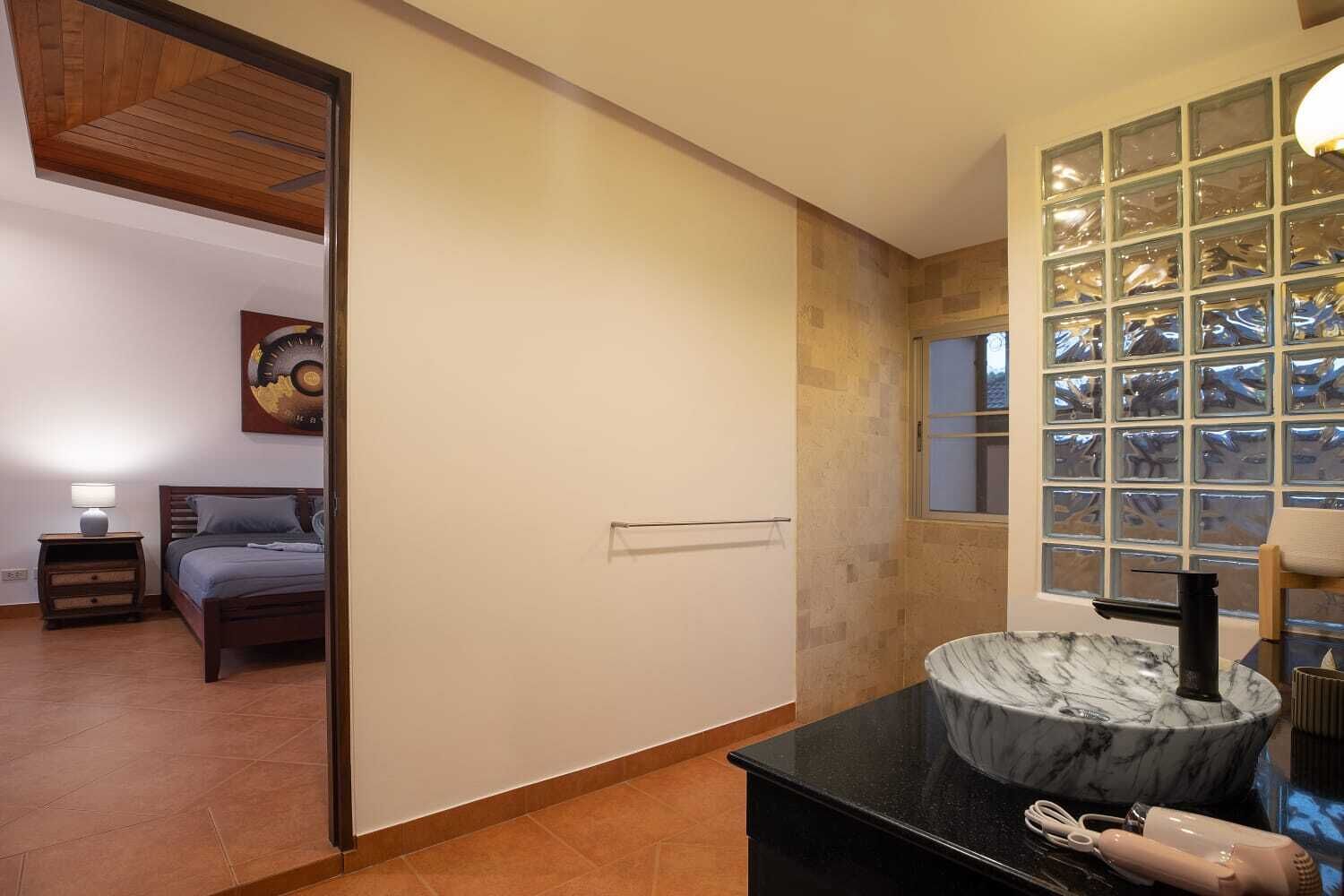 Un dormitorio con cama, mesita de noche, lámpara y vista a otra habitación por una puerta abierta.