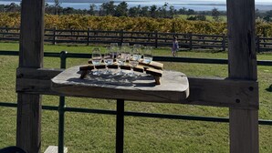 Une enseigne en bois, sur laquelle est fixée une plaque métallique portant l’inscription « The Gurdies Winery », et sur laquelle sont perchés deux oiseaux. Devant, on aperçoit une table avec des verres à vin vides et une planche en bois.