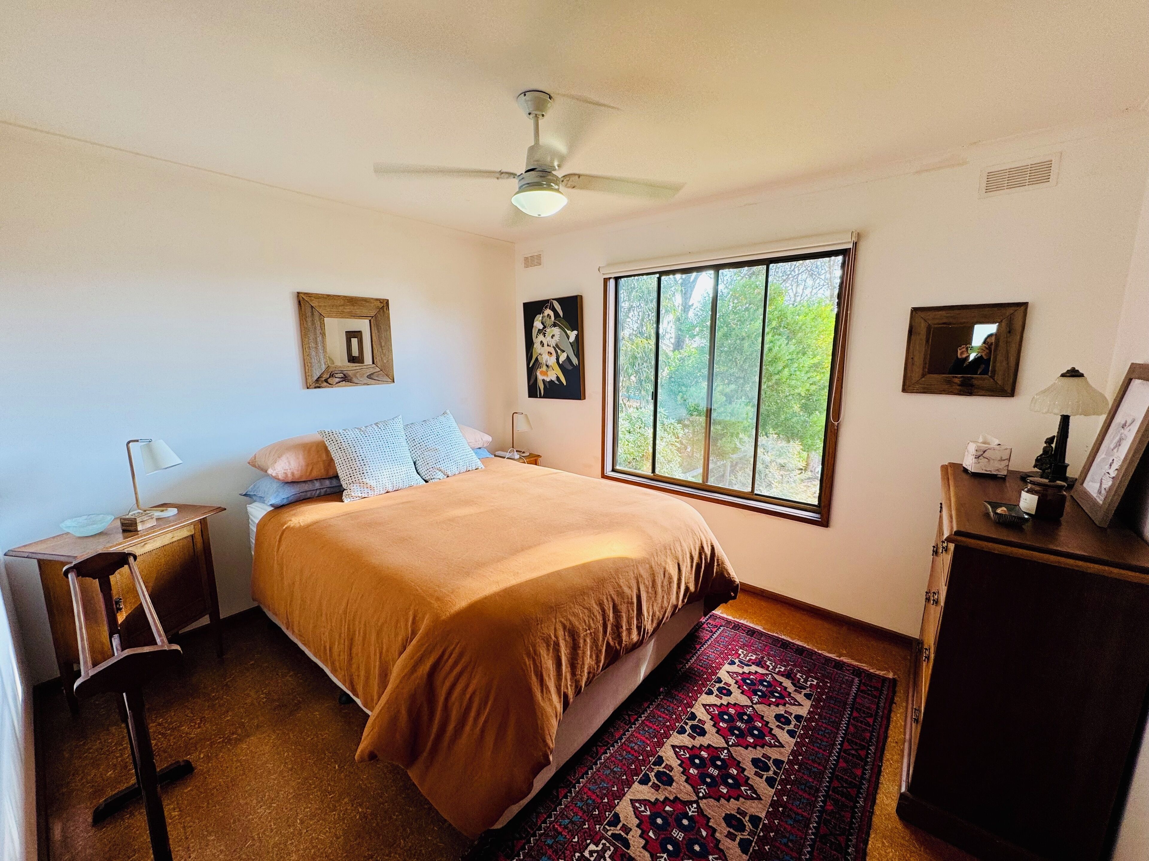 Une chambre avec une grande fenêtre, un ventilateur de plafond, un lit recouvert d’une couverture marron, une table de chevet avec une lampe et une commode avec un miroir et un cadre photo.