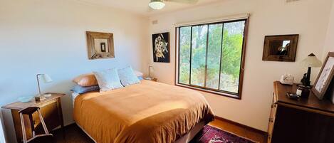 Une chambre avec une grande fenêtre, un ventilateur de plafond, un lit recouvert d’une couverture marron, une table de chevet avec une lampe et une commode avec un miroir et un cadre photo.