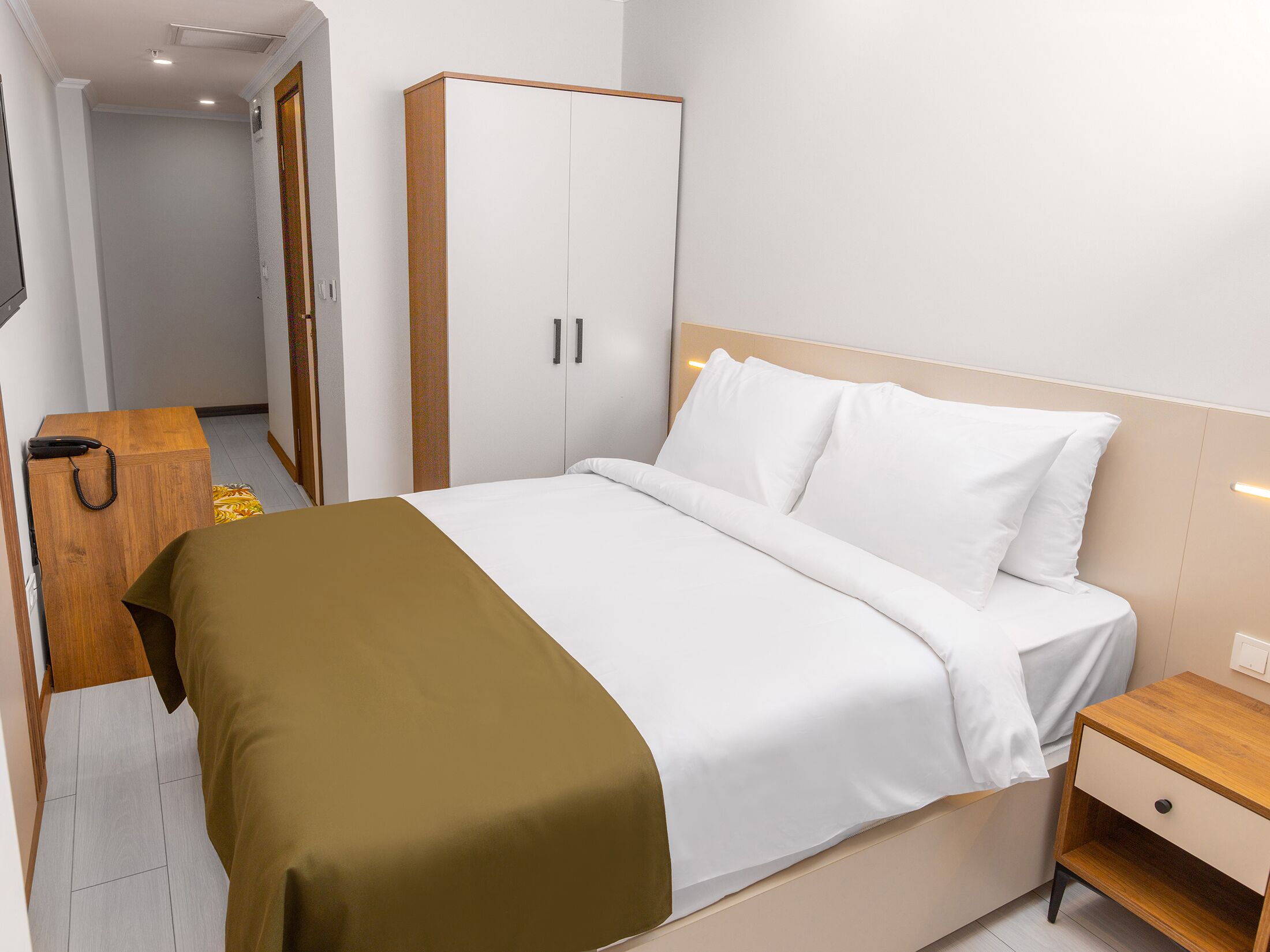 Een hotelkamer met een bed, een nachtkastje van hout, een kledingkast en een telefoon op een houten tafel.