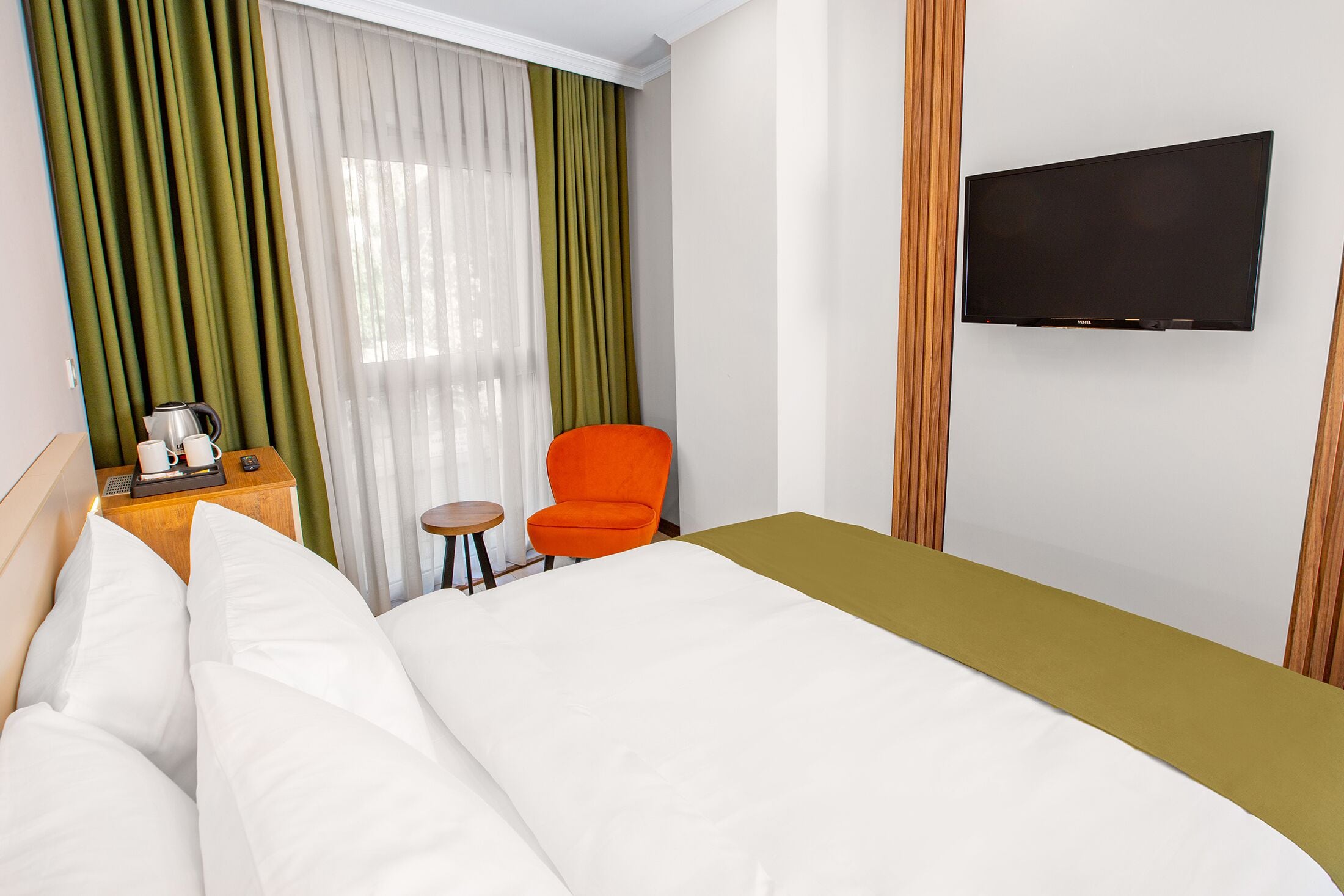 A hotel room with a bed, a television mounted on the wall, a small table with a kettle and cups, and an orange chair.