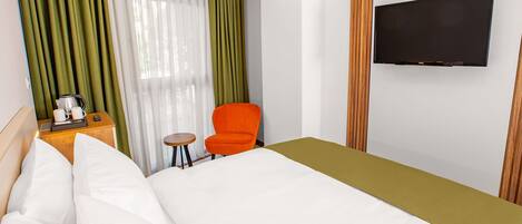 A hotel room with a bed, a television mounted on the wall, a small table with a kettle and cups, and an orange chair.