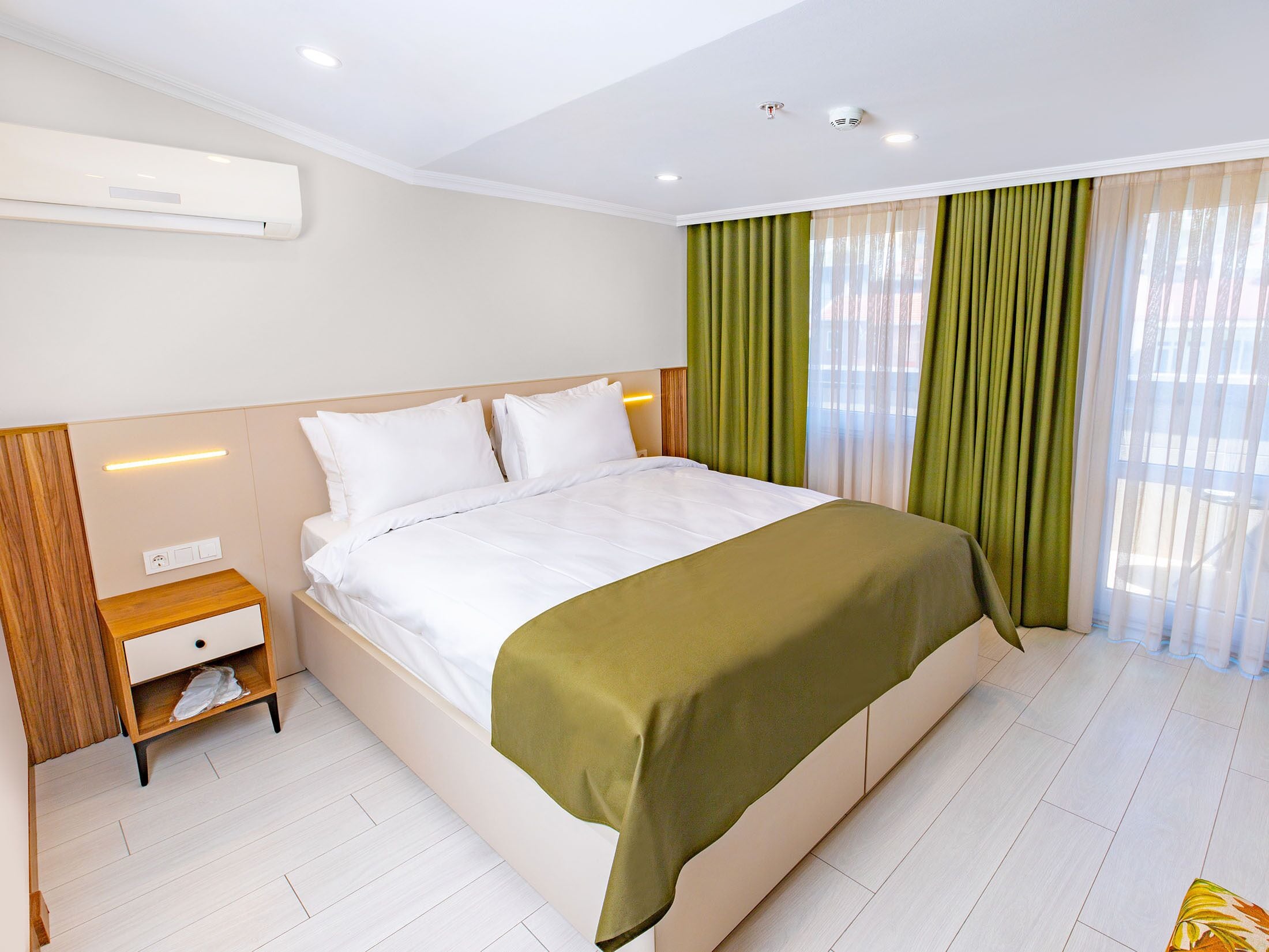 Une chambre d’hôtel moderne dotée d’un grand lit, d’une table de chevet, d’un climatiseur intégré et d’une fenêtre avec des rideaux légers.