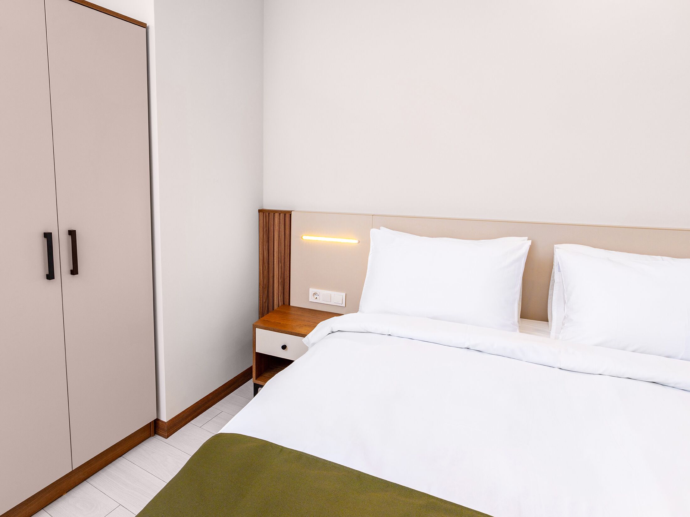 A hotel room with a bed, a nightstand, a wooden wardrobe, and a wall-mounted light.