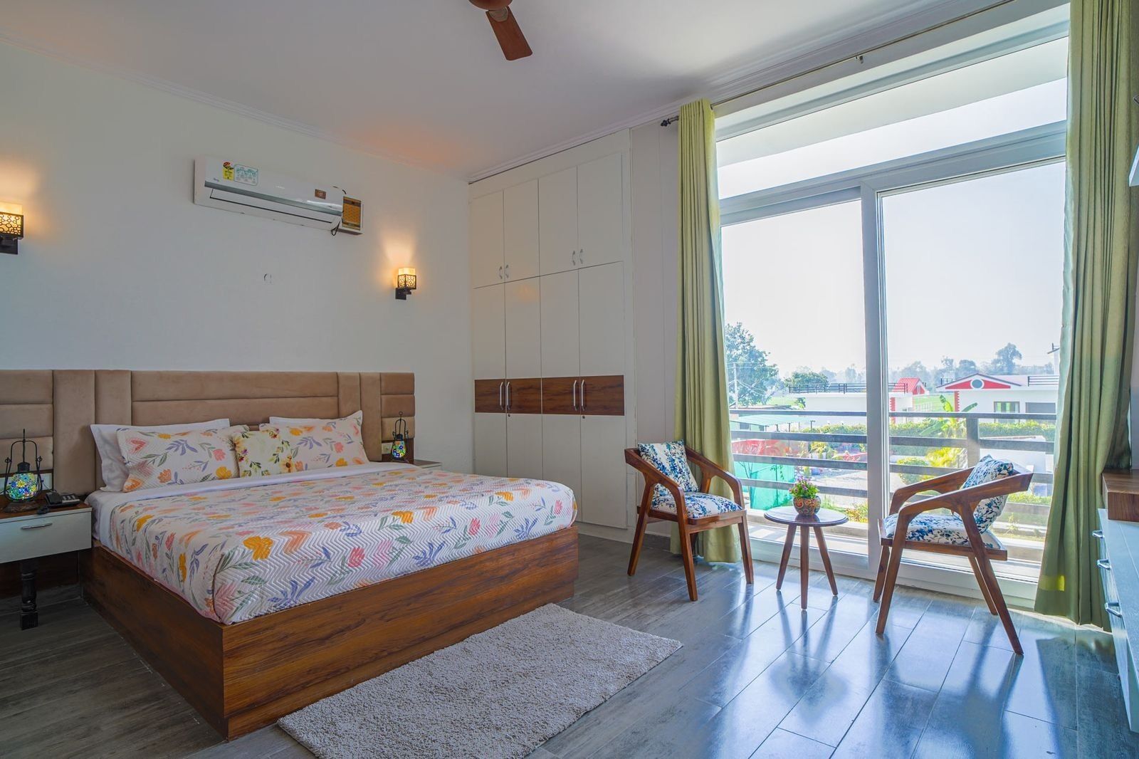A bedroom with a large bed, a wardrobe, a nightstand, two chairs, a small table, and a large window with a view of buildings.