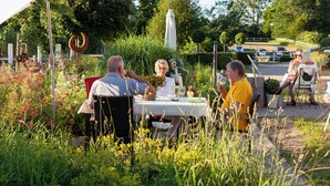 A group of people dining outdoors at a table with a white tablecloth, surrounded by lush greenery and flowers. - Hof Eckhorst - View apartment - Stylish ambience & cozy atmosphere (Güby)