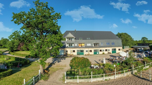 A large building with multiple windows and a green roof, surrounded by a fenced area with a garden and a sandy path. - Hof Eckhorst - View apartment - Stylish ambience & cozy atmosphere (Güby)