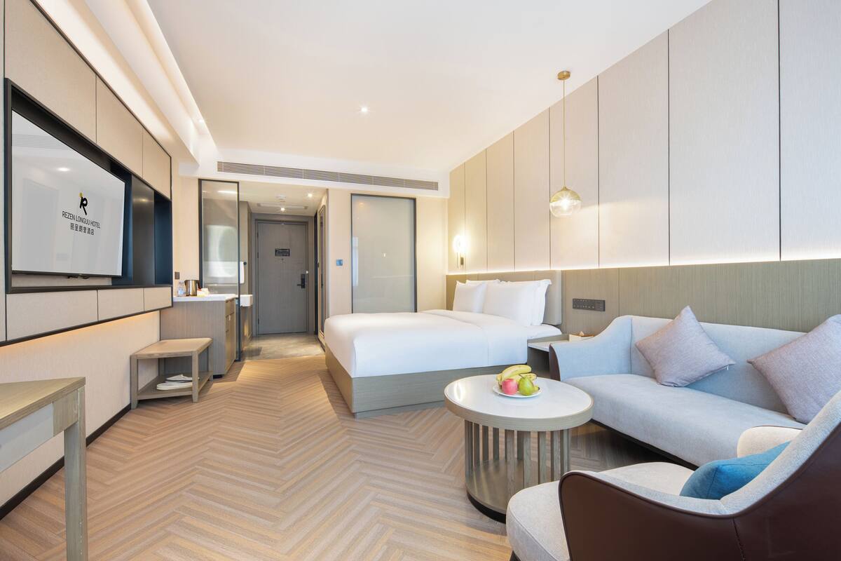 A modern hotel room with a bed, a sofa, a TV, and a small table with fruit.