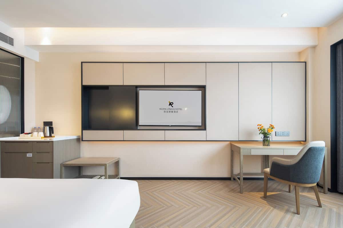 A hotel room with a bed, a desk, a chair, a TV mounted on the wall, and a small table with a vase of flowers.
