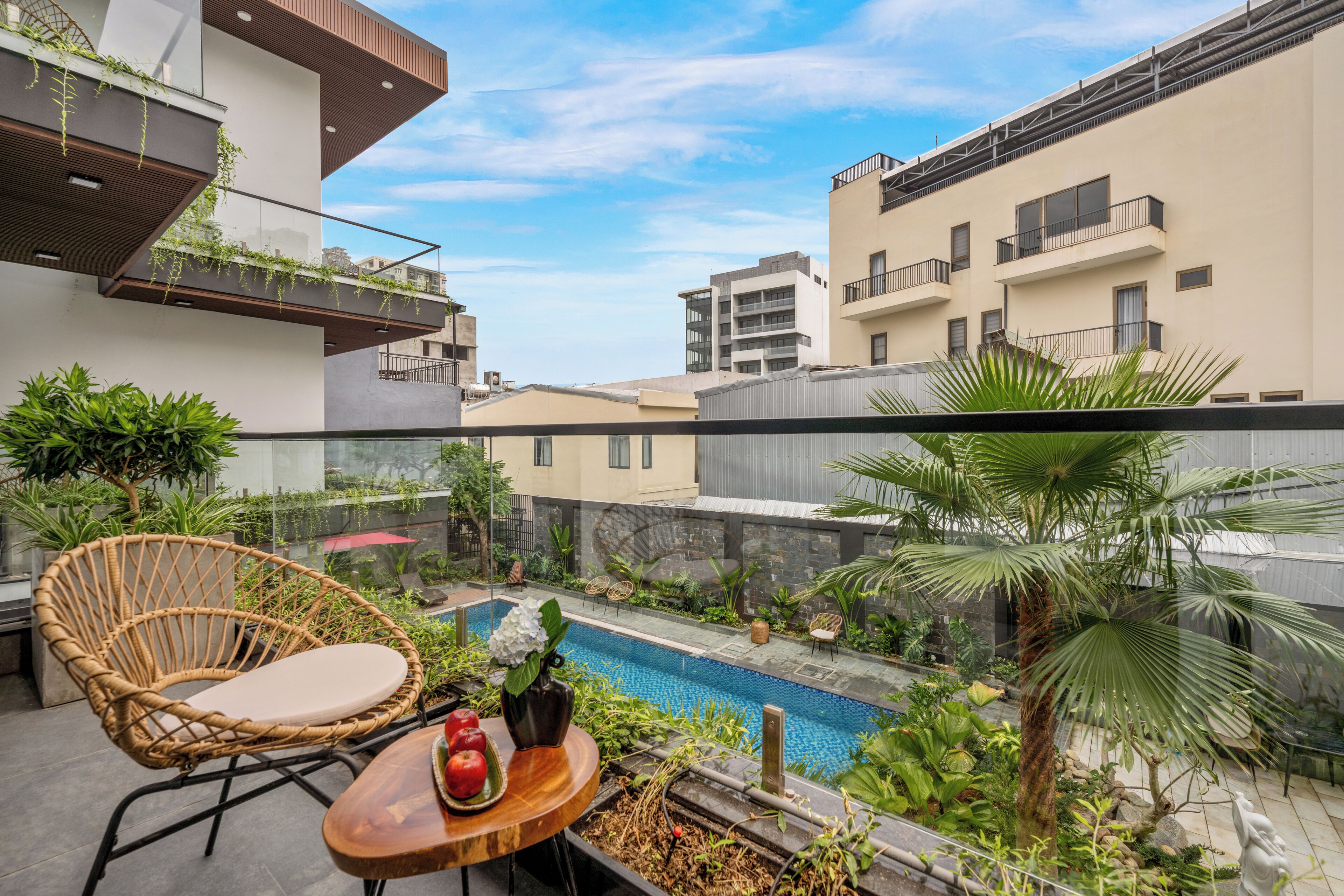 A balcony with a wicker chair, a small table with apples and a vase, and a view of a swimming pool and buildings in the background.