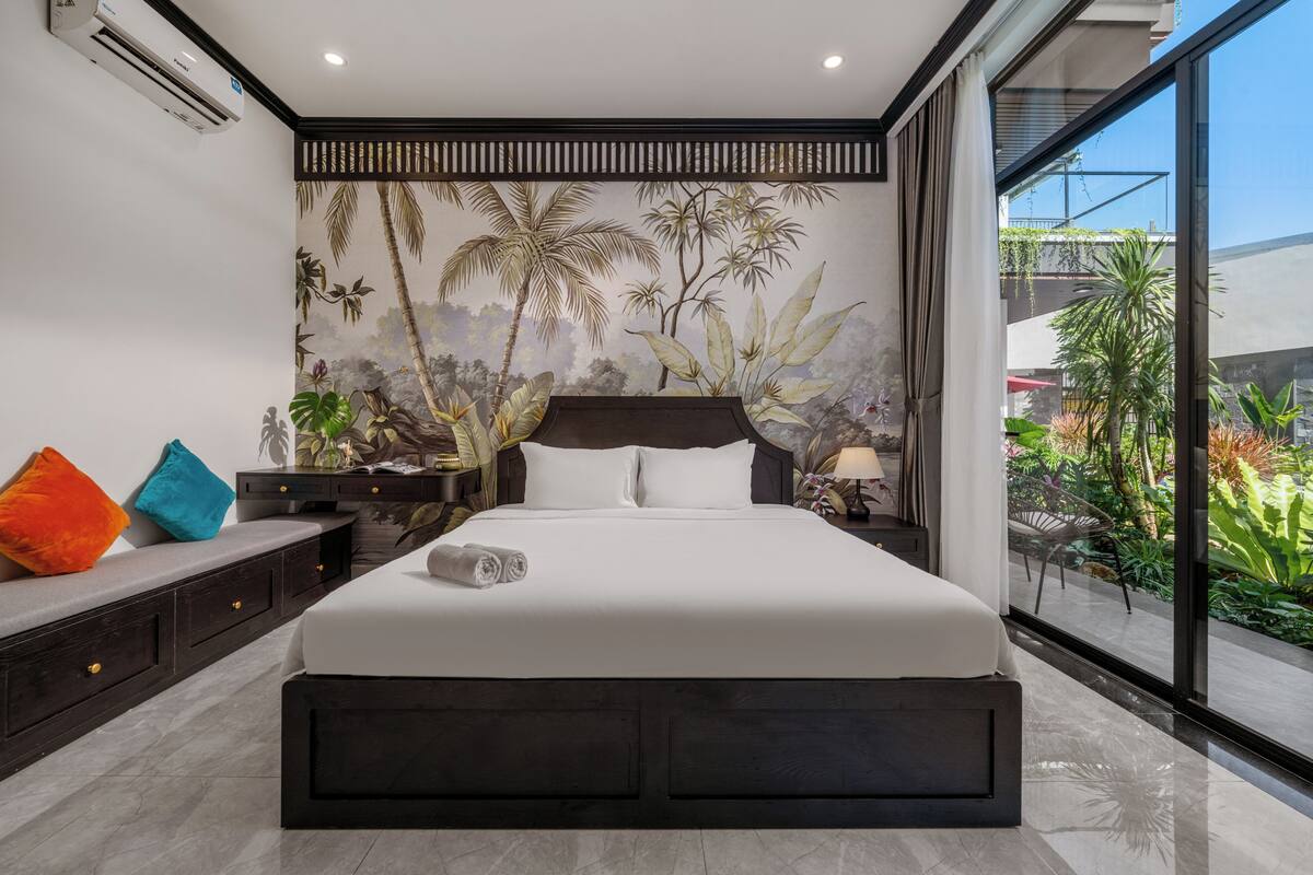 Une chambre moderne avec un grand lit, une fresque représentant une végétation tropicale, une vue sur la verdure à l’extérieur et une porte-fenêtre coulissante.