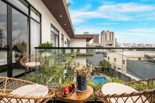 A balcony with wicker chairs, a wooden table with fruit and flowers, and a view of a cityscape and a swimming pool. - CiCi Villa & Apartment (Da Nang)