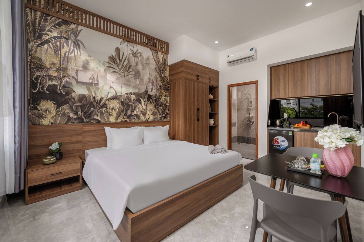 Une chambre d’hôtel moderne dotée d’un grand lit, d’une tête de lit en bois, d’une table à manger avec des chaises, d’un coin cuisine et d’une grande fresque murale.