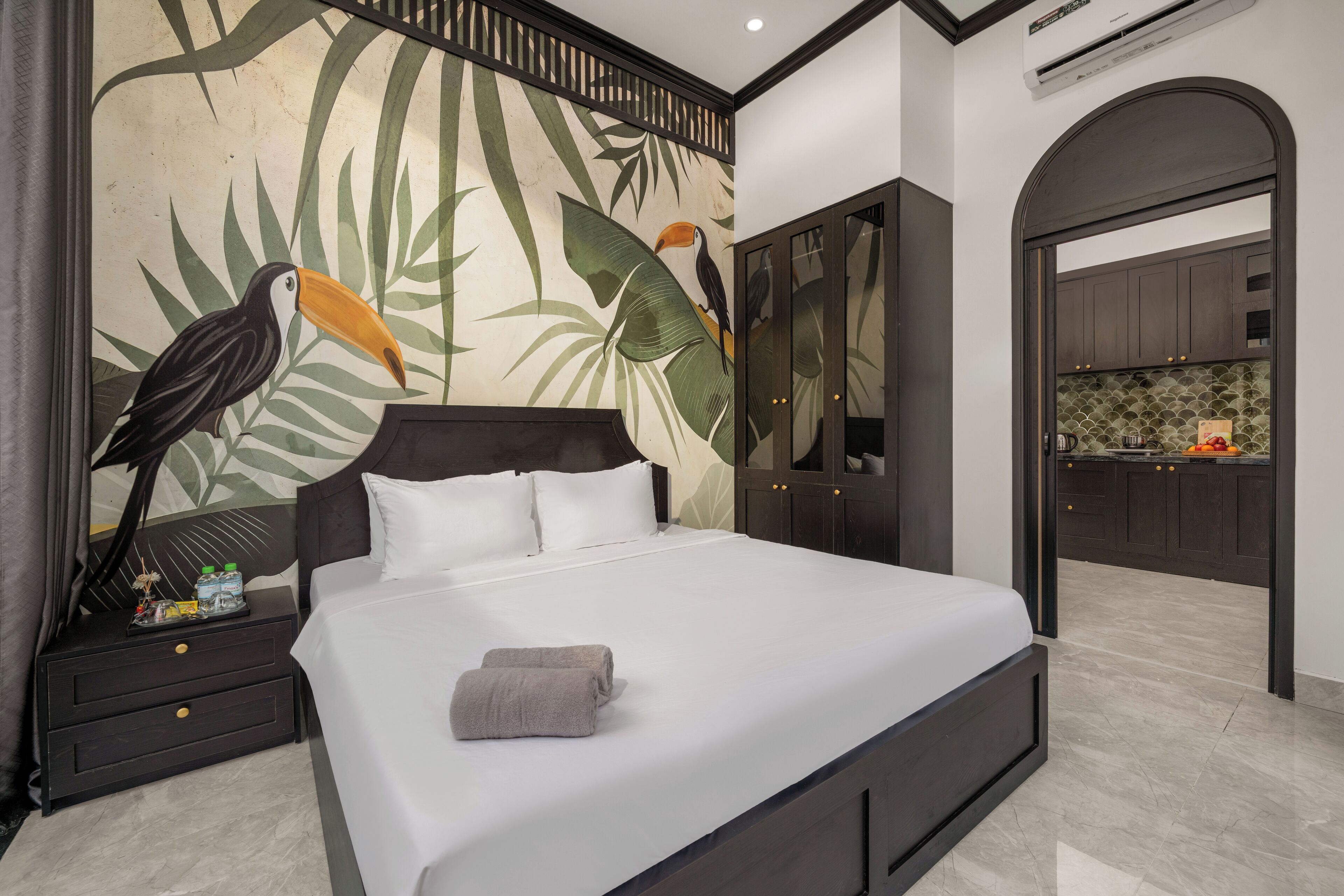 A modern bedroom with a large bed, a nightstand with toiletries, a mural of toucans, and a view into the kitchen.