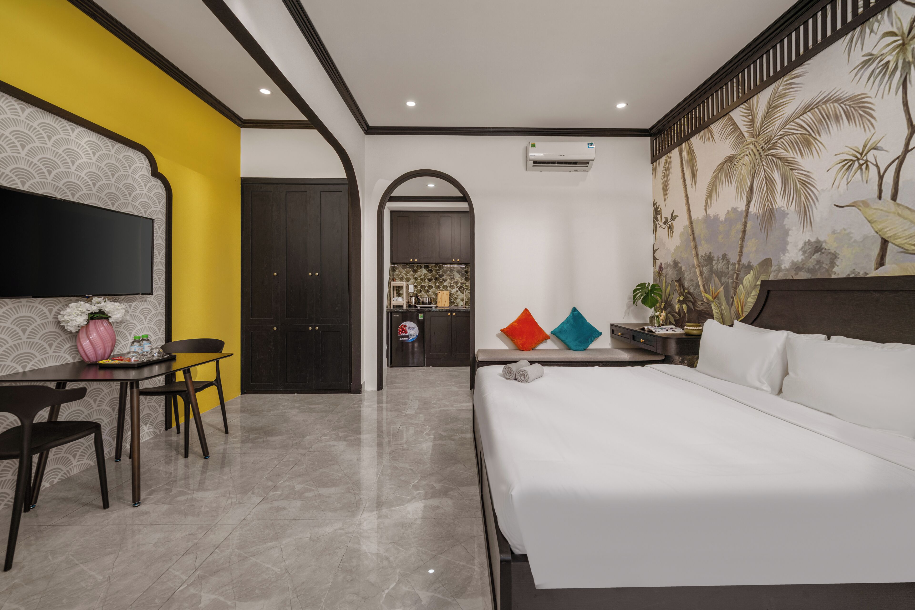 A hotel room with a bed, a TV, a dining area, and a kitchen in the background.