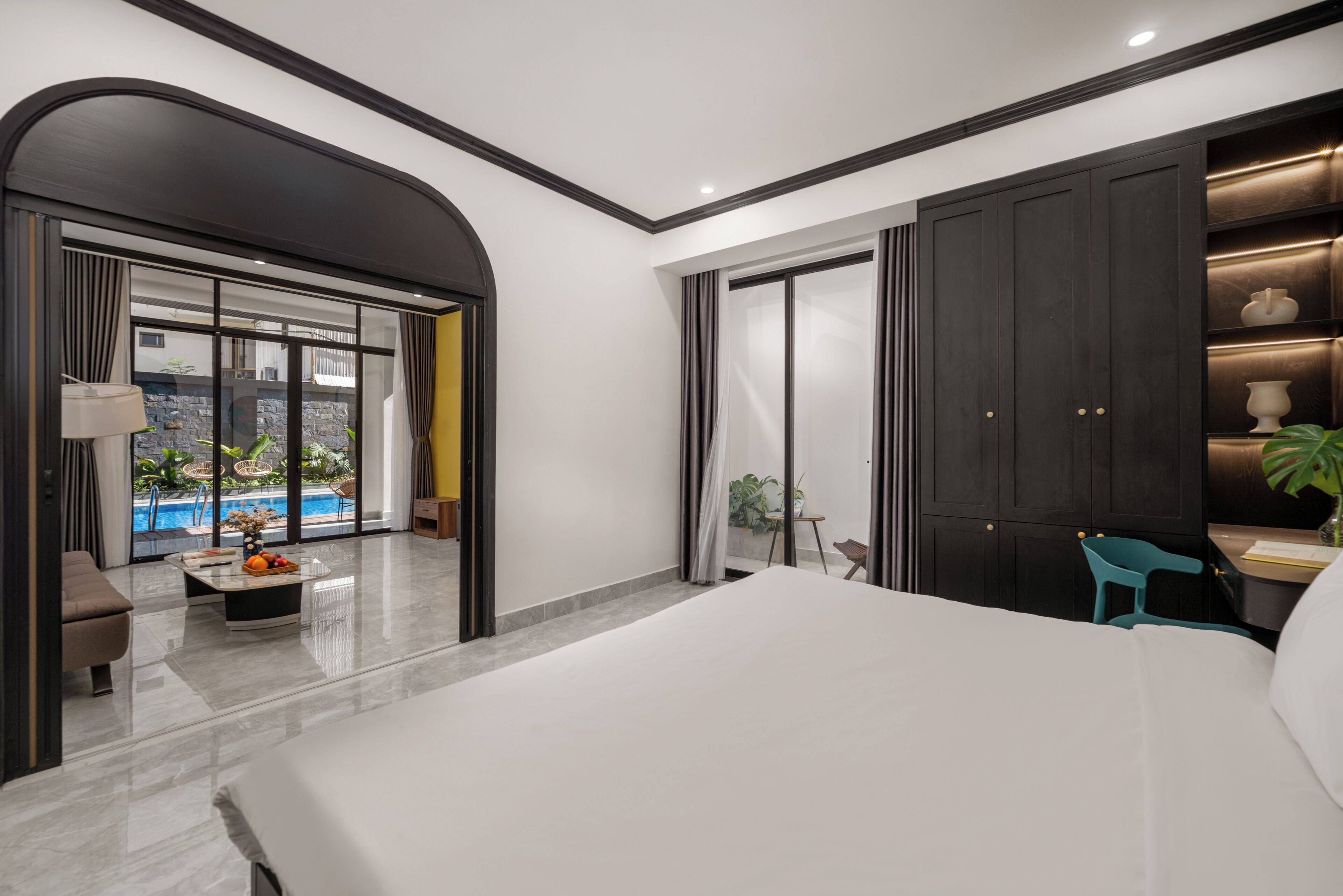 A modern bedroom with a large bed, a desk with a chair, a view of a swimming pool, and a dark wardrobe.