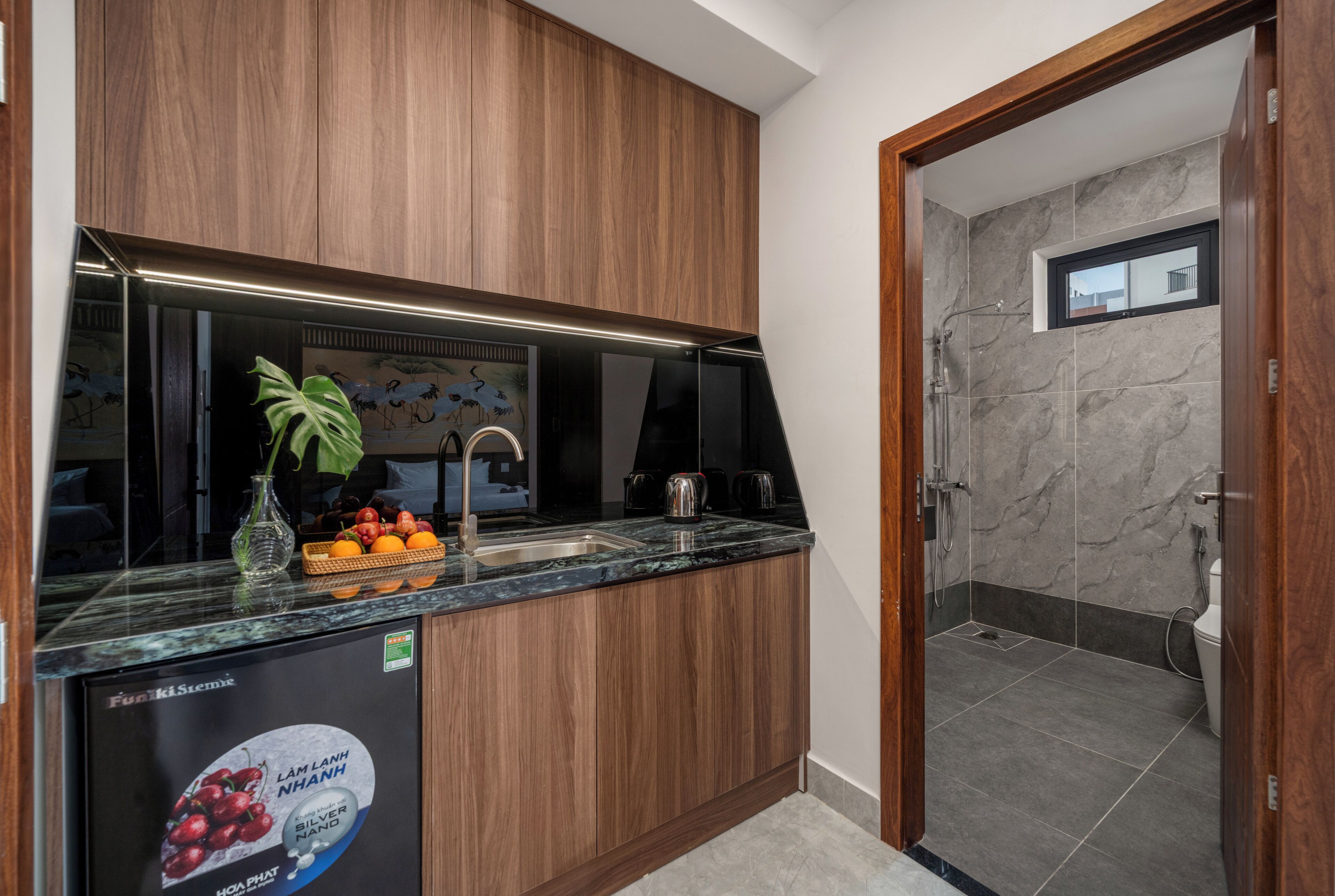 A modern kitchen with wooden cabinets, a marble countertop, and a sink. There is a small window in the adjacent bathroom.