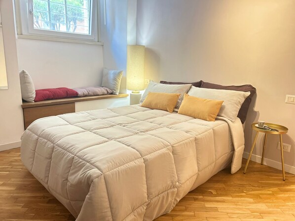 A neatly made bed with a beige duvet, a bedside table with a lamp, and a window with a view of greenery. - SAINT JOHN HOUSE ROME only 10 minutes by subway from the historic center of Rome (Roma)