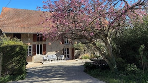 A stone building with a tiled roof, a gravel driveway, and a garden area with a bench and hanging flower baskets. - Gîte le Haut des Lariers (Morlet)