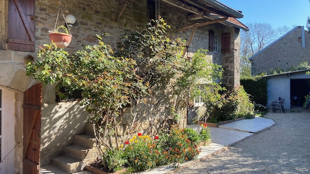 A stone building with a wooden staircase leading to a door, surrounded by a gravel driveway and a small garden with flowers. - Gîte le Haut des Lariers (Morlet)