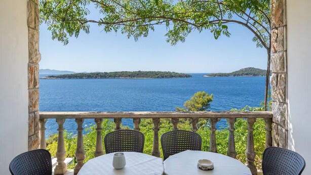 A balcony with a table and chairs overlooking a clear blue sea and distant islands. - 3-Bedroom Apartment in Prizba with Ocean Views - Ideal for Families and groups (Smokvica)