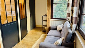 Une pièce chaleureuse avec un canapé gris, un plancher en bois, une fenêtre avec des rideaux et une porte dotée d’un cadre noir.
