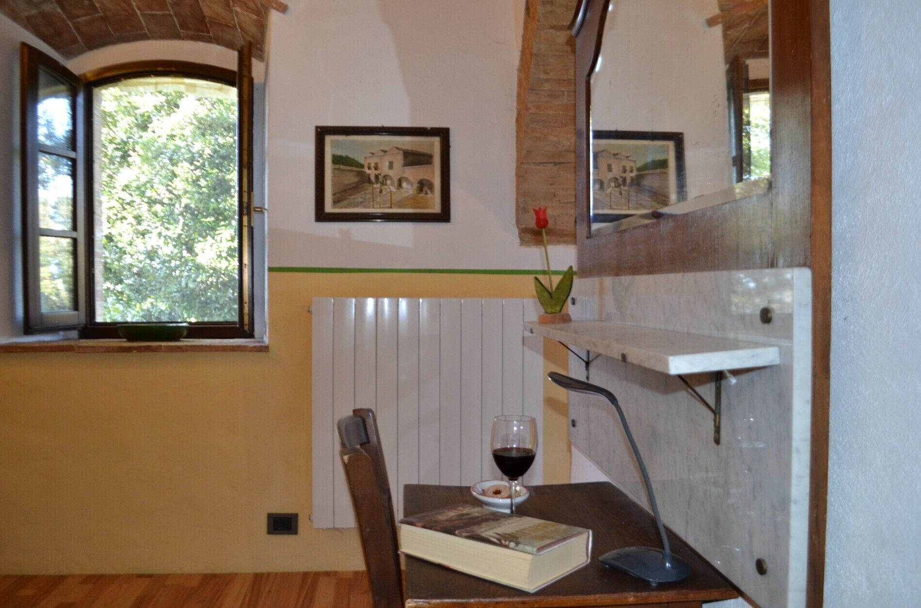 A room with a wooden floor, a dining table with a book and a glass of red wine, a chair, a radiator, and a window with a view of trees.